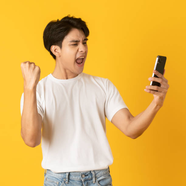 Young good looking asian man shouting happily on smartphone isolated on yellow background stock photo