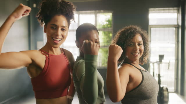 Portrait of strong young women flexing their biceps in a gym. Diverse group of powerful, confident and empowered athletic female friends smiling and feeling proud of their toned muscles from training