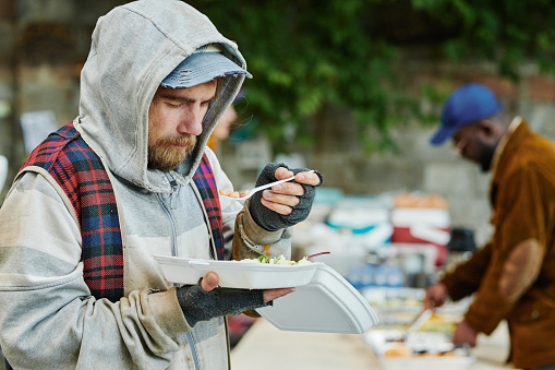 Homeless man in warm torn clothing eating food outdoors giving by volunteers during charity
