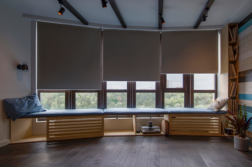 Motorized roller shades in the interior. Automatic roller blinds blackout fabric on large windows. Remote Control Shades are above the windosill with pillows. Summer. Green trees outside.