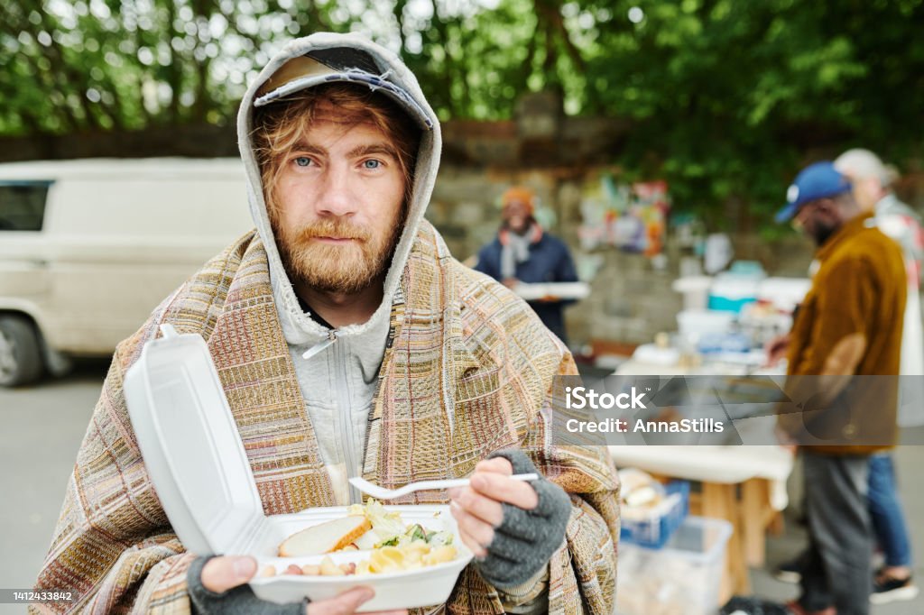 Homeless man eating food outdoors Portrait of bearded homeless man in warm clothing looking at camera while eating food outdoors during charity Homelessness Stock Photo