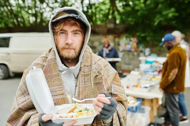 Portrait of bearded homeless man in warm clothing looking at camera while eating food outdoors during charity