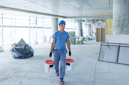A young Caucasian male construction worker wearing a hardhat is carrying buckets of paint through a demolished room.