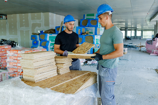 Two young Caucasian male construction workers wearing protective equipment are standing in a demolished room and making piles of wooden floor tiles.