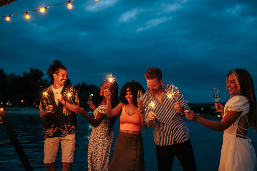 A group of young people are partying at the riverside with drinks and sparklers on a warm summer night.