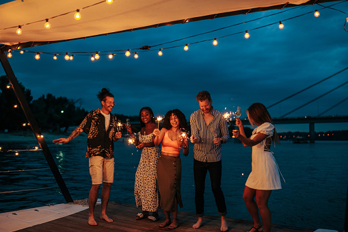 Young and beautiful multiracial friends enjoy partying on the boat with sprinklers and drinks. Summer evening on the river with the dark blue background