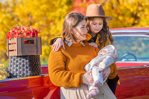 full-bodied mother with two little daughters stands near a red retro car loaded with boxes of vegetables against the backdrop of rural autumn nature.