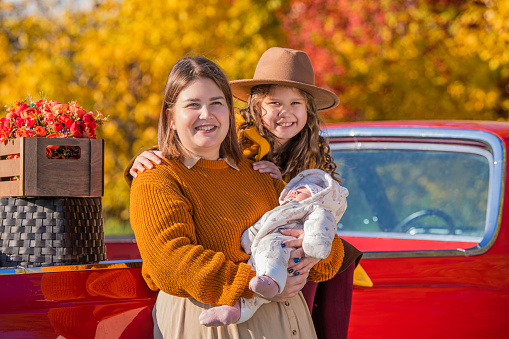 full-bodied mother with two little daughters stands near a red retro car loaded with boxes of vegetables against the backdrop of rural autumn nature.