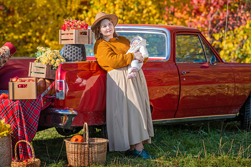 full-bodied mother with a newborn little daughter stands near a red retro car loaded with boxes of vegetables against the backdrop of rural autumn nature.