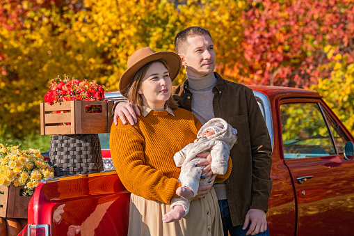 full-bodied mother with a newborn little daughter and father stands near a red retro car loaded with boxes of vegetables against the backdrop of rural autumn nature.