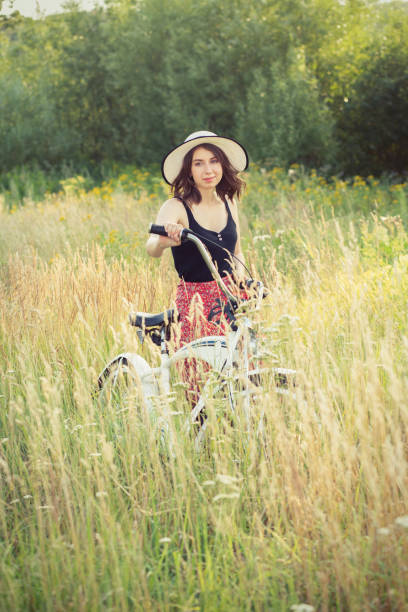 happy brunette girl in hat walking with bicycle at sunny meadow, looking at camera