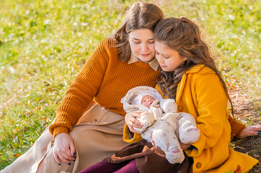 happy full-bodied mother sits on the grass next to her older daughter hugging her newborn sister in the autumn park on a sunny day.
