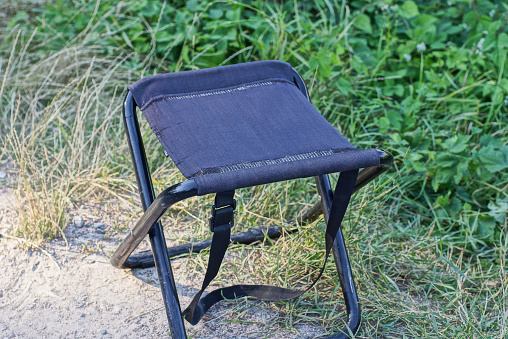 one black folding chair made of fabric and metal stands on gray ground in green grass and vegetation in nature