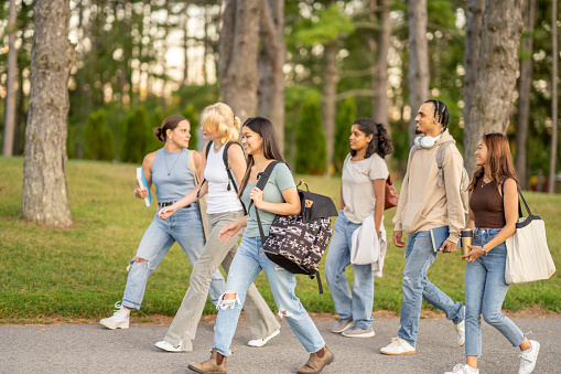 A group of University students are seen walking outside on campus as they make their way to class.  They are dressed casually and have backpacks on as they talk together and make their way to school for the day.