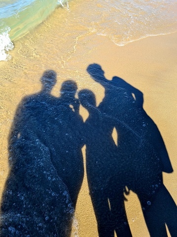 Shadow of a family on the sand. The waves and foam lick the shadow of four people. We see the shadow of the person taking the picture.
