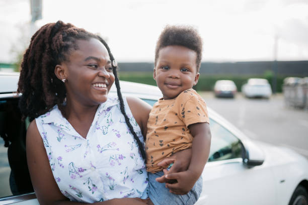 Portrait of a little baby boy held by his mother and looking at the camera while she is standing next to her electric vehicle parked by the charging station