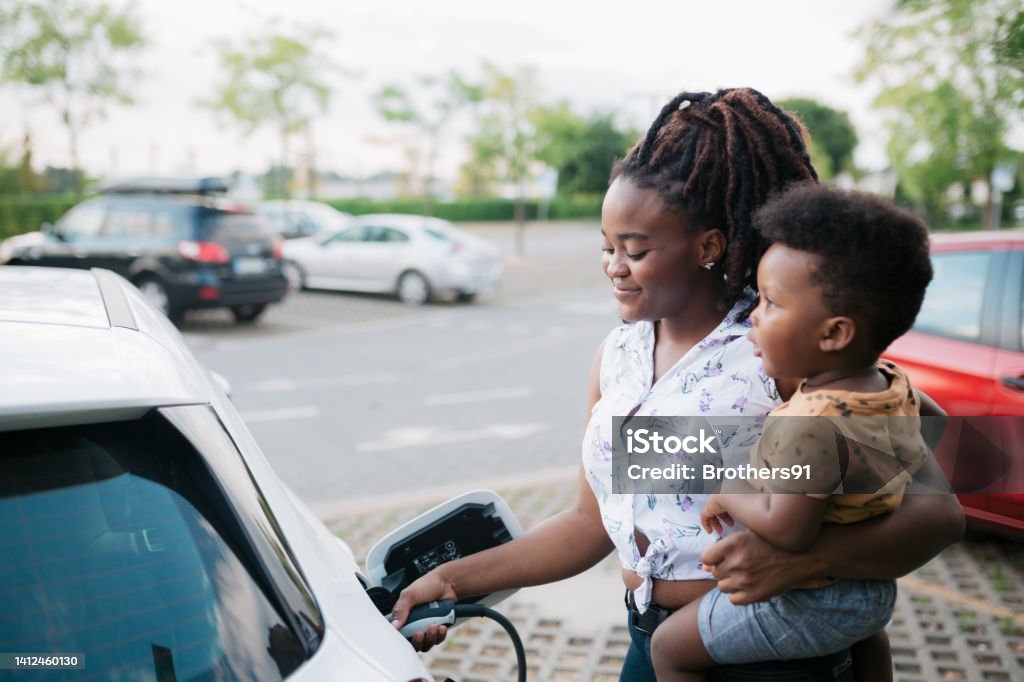 Mother holding her son while charging her electric vehicle Young adult mother charging her electric car parked next to the charging station and holding her baby son in her other hand Electric Vehicle Stock Photo