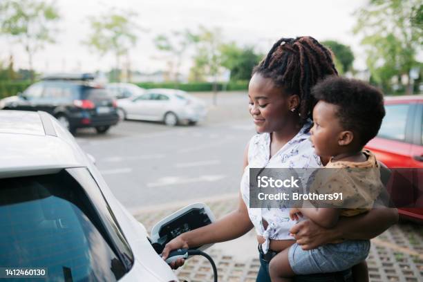 Mother Holding Her Son While Charging Her Electric Vehicle Stock Photo - Download Image Now