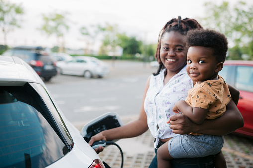 Portrait of a happy mother charging her electric car parked next to the charging station and holding her baby son in her other hand, both of them looking at the camera