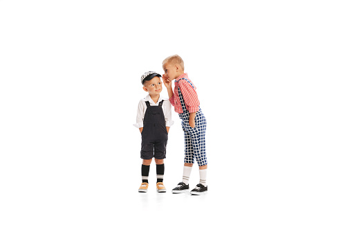 Portrait of two little boys, children in stylish retro costumes posing isolated over white studio background. Whispering secrets. Concept of childhood, friendship, fun, lifestyle, fashion. Retro style