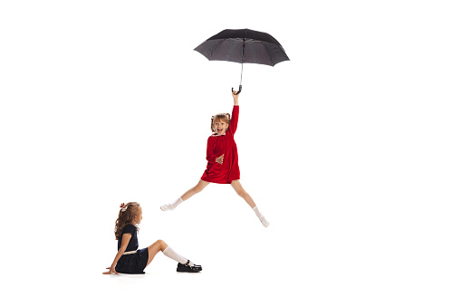 Portrait of two little girls, children playing together with umbrella isolated over white studio background. Flying. Concept of childhood, friendship, fun, lifestyle, fashion. Retro style