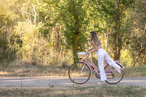 Side view of young elegant woman getting on retro pink bike in the middle of nature in spring time in the village