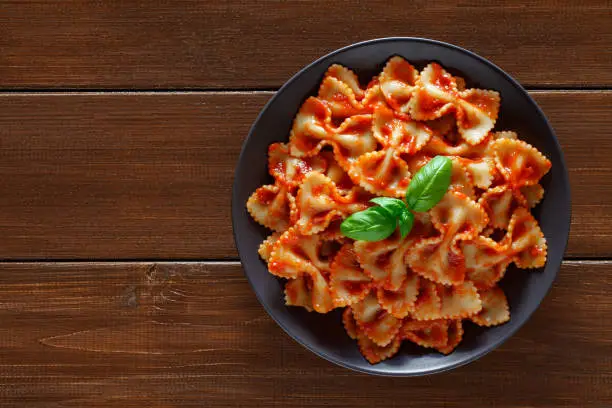 Farfalle, cooked pasta with tomato sauce and basil green leaf, in gray plate, on wooden brown plank tabletop background, top view, space to copy text.