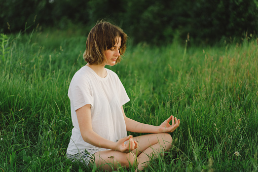 Beautiful young girl in a white T-shirt  doing yoga in a beautiful field. Healthy lifestyle and mindfulness concept