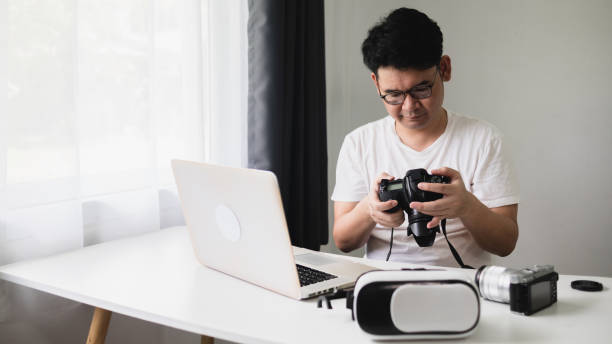 istock Content creator concept In virtual reality, an Asian man is looking down at a camera to edit on a laptop computer in his living room at home. 1412481154