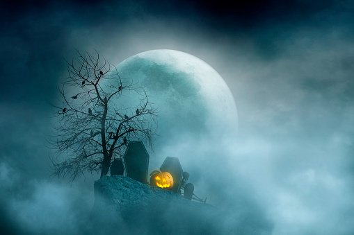 An illuminated jack o'lantern rest in a small cemetery on a hill in front of a large rising moon on a spooky Halloween night.