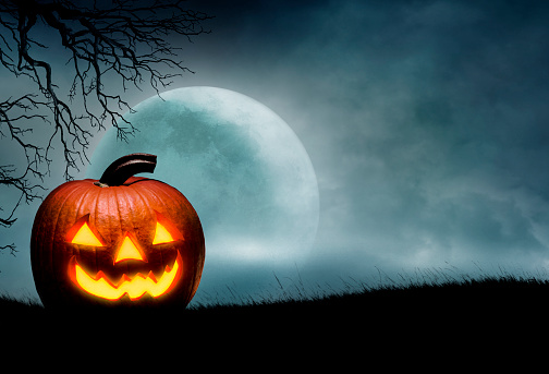 An illuminated jack o'lantern rests on a grassy hill and a rising full moon.