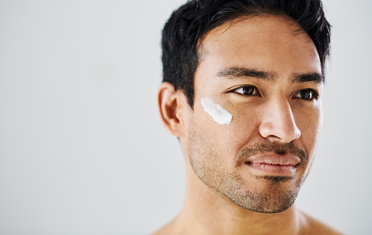 Man applying cream moisturizer to his face while grooming against a grey copy space background. Handsome young guy using sunscreen lotion with spf for uv protection, healthy complexion and clear skin