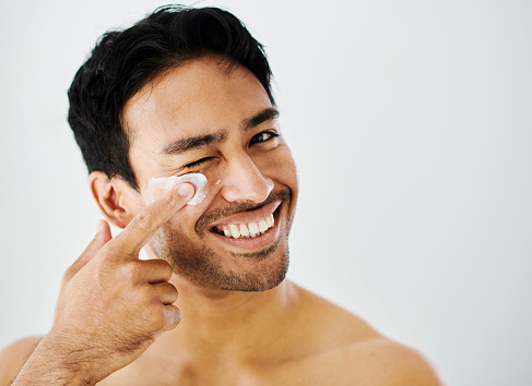 Skin and beauty routine for a man with lotion or moisturizer against a white studio background. Portrait of a happy, smiling and confident male applying cream or sunscreen to his face for protection