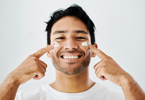 Face, beauty and skincare routine of a handsome young man applying moisturizer with his finger. Confident male smiling in studio, isolated against a grey background during his antiaging regimen