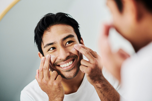 Face, beauty and skincare treatment of a handsome man applying moisturizer with his fingers. Confident male smiling and looking at his reflection in the bathroom mirror during his antiaging regimen