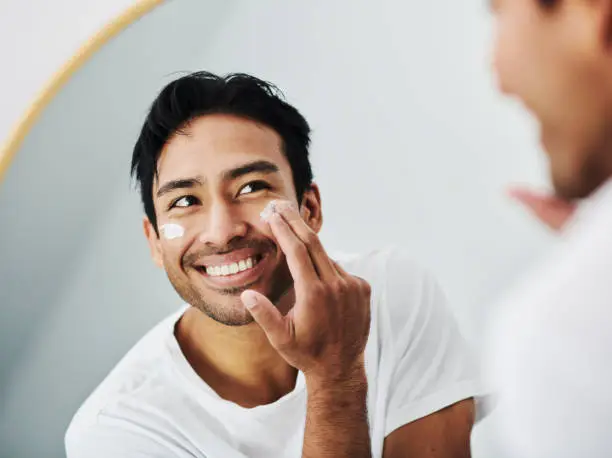 Photo of Man applying face skincare cream while grooming looking at the mirror in the bathroom in his morning routine at home. Close up portrait of handsome guy using facial lotion for clean and clear skin