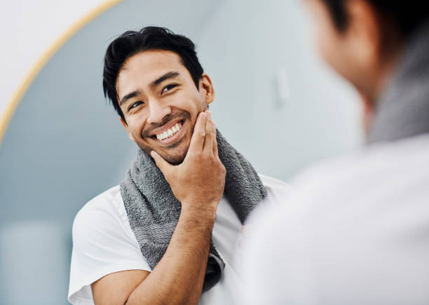 toelettatura, igiene e sana routine di cura della pelle del viso per una pelle maschile liscia e morbida. bel giovane che si guarda allo specchio toccandosi la barba felice del suo prodotto per la rasatura facciale - shorn foto e immagini stock