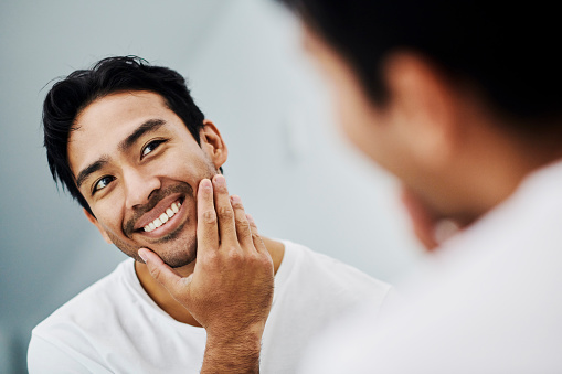 Handsome male touching his healthy face in the mirror after a skincare treatment. Confident man feeling fresh and attractive from a beauty routine. Portrait of a guy looking at his skin.