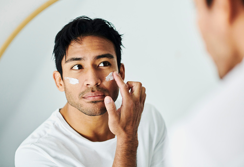 Man applying cream, lotion and moisturizer for a skincare routine while grooming in a mirror at home. Handsome young guy using sunscreen lotion with spf for uv protection on his face for healthy skin