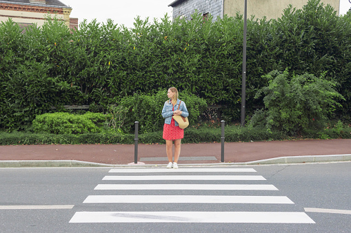A girl in a red dress and a denim jacket with a bag on her shoulder stands at a pedestrian crossing and looks to cross the road