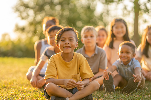 A large group of school-aged children sit together on the grass as they pose for a portrait.  They are each dressed casually in t-shirts and shorts as they smile and enjoy the time together.  The focus is on a sweet boy of Asian decent in front who is smiling.