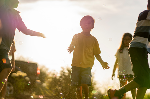 A small group of children are seen playing with bubbles as they try to pop them with their hands.  They are each dressed casually and are smiling as they run around in the summer sun giggling and laughing.