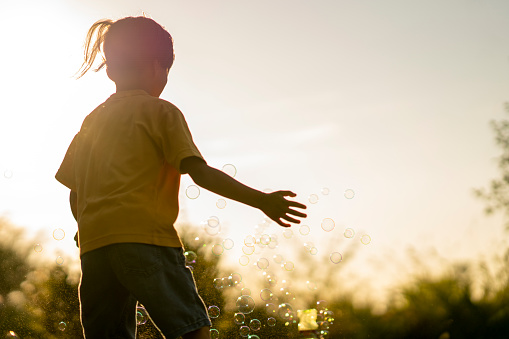 A sweet little boy of Asian decent smiles as he plays outside on a hot summers day.  He is dressed casually and is trying to pop bubbles as he smiles and laughs in the sunlight.