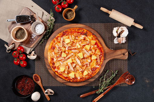 Hawaiian Pizza with raw cherry tomato, black pepper, garlic, and mushroom isolated on wooden cutting board on dark background top view of fastfood