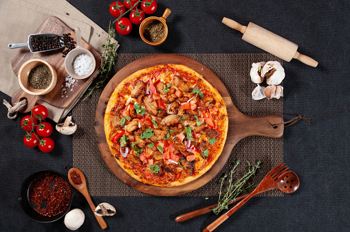 Butter Chicken Pizza with raw cherry tomato, black pepper, garlic, and mushroom isolated on wooden cutting board on dark background top view of fastfood