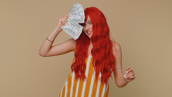 Redhead young woman holding fan of cash money dollar banknotes celebrate dance, success business career, lottery game winner, big income, wealth. Red hair ginger girl alone on beige wall background