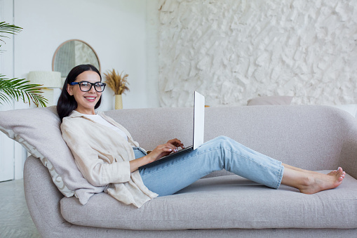 Portrait of young beautiful woman in glasses working at home sitting on sofa and looking at camera smiling, brunette uses laptop for remote homework and study