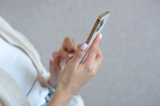 A close-up photo of a beautiful woman's hand with a white manicure is using a gold-colored phone, typing a message on a smartphone