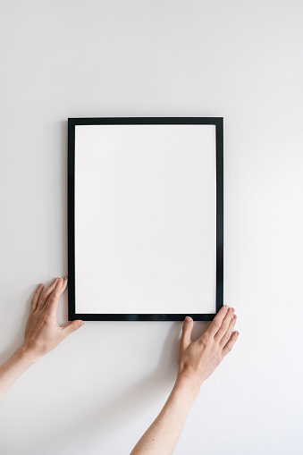 Cropped vertical view of woman hand install copy space picture frame. Female hold blank poster against white wall. Art placard hanging in room. Concept of home decor