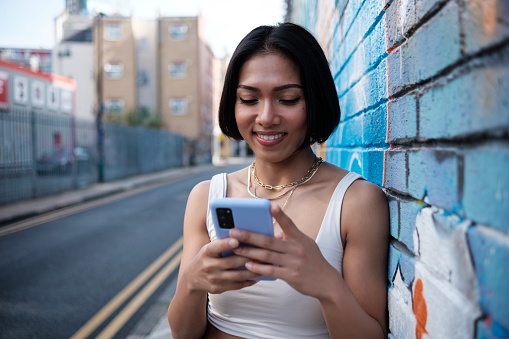 Young asian woman leaning against a colorful wall and using her phone. She is smiling and wearing a sleeveless white t-shirt.